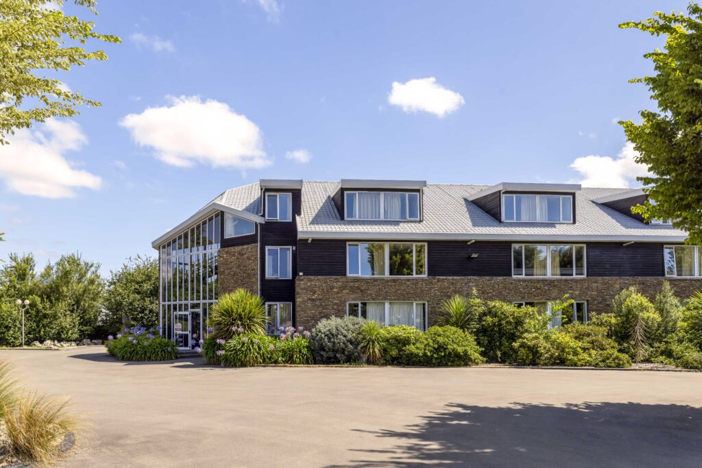 Exterior view of Methven Resort hotel with stone and timber facade, landscaped gardens and clear blue sky.