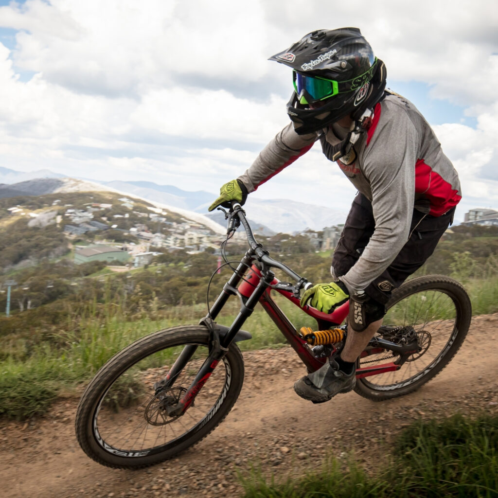 Mountain biker wearing helmet and goggles riding downhill trail with scenic mountain views in the background.