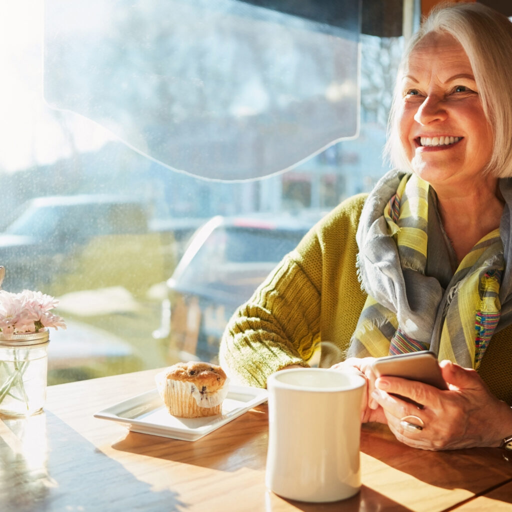 Smiling senior woman enjoying coffee and muffin in a sunny café, holding smartphone by window.