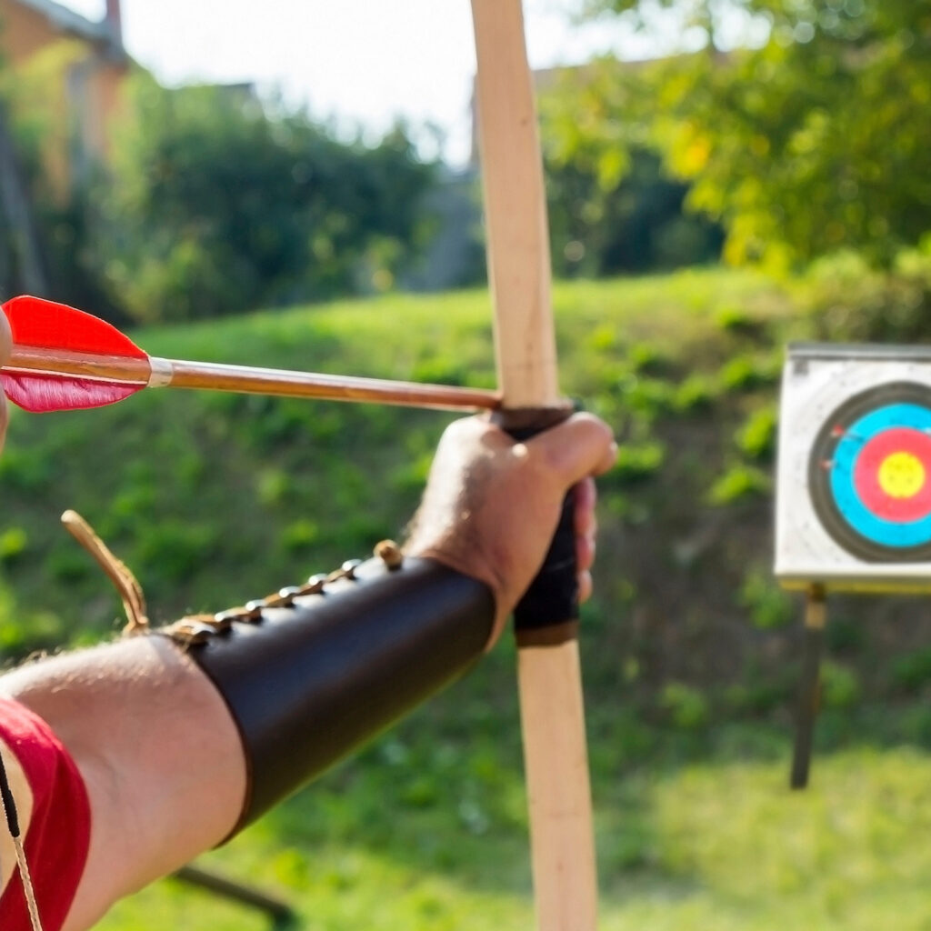 Archer aiming wooden bow with red arrow at outdoor target, archery practice in sunlight.