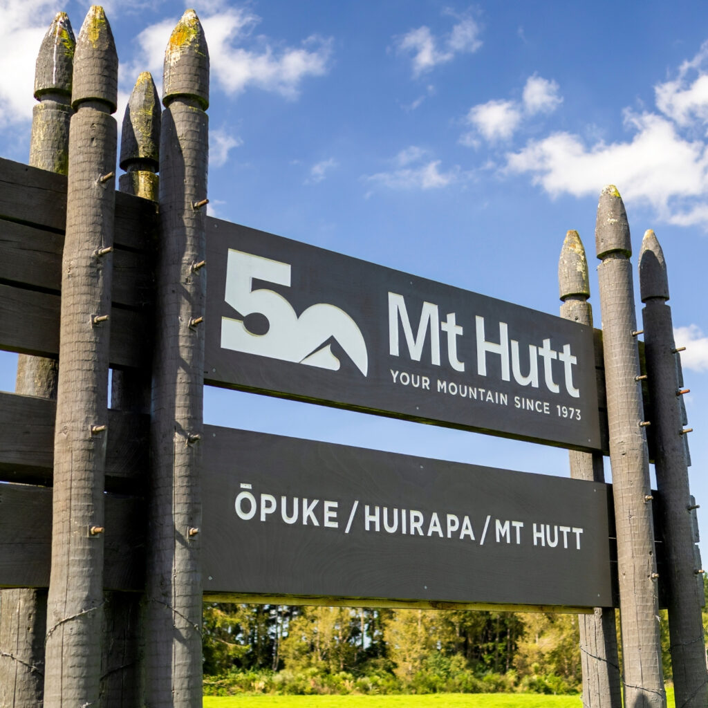 Mt Hutt ski field entrance sign with 50th anniversary logo and bilingual text Ōpuke / Huirapa / Mt Hutt under blue sky, Canterbury New Zealand.