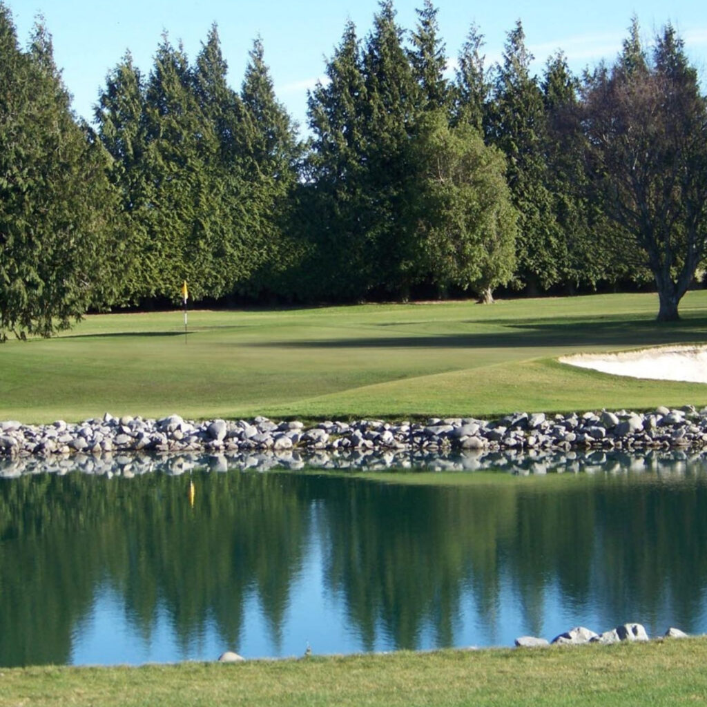 Golf course green with flagstick, sand bunker and pond reflecting trees on a sunny day.