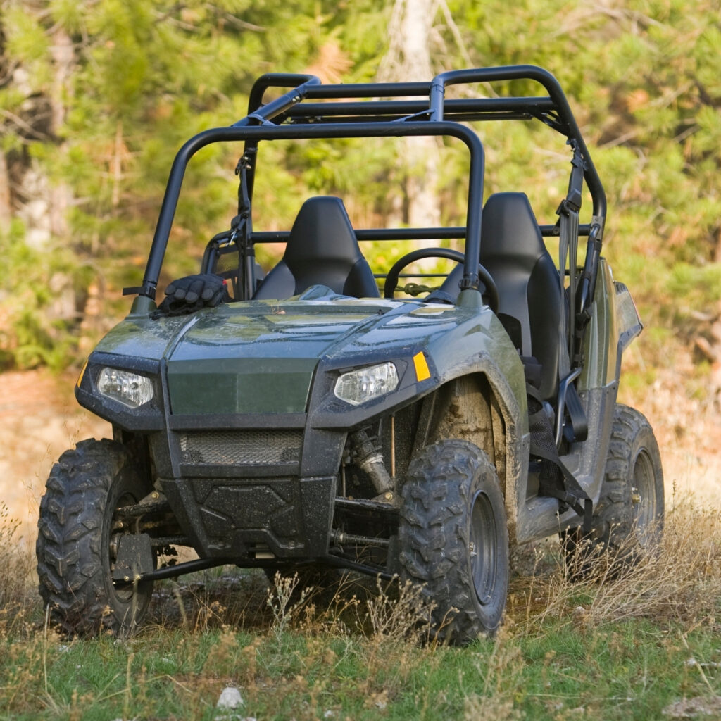 All-terrain utility vehicle (UTV) parked outdoors on grassy trail with roll cage and off-road tires, forest background.