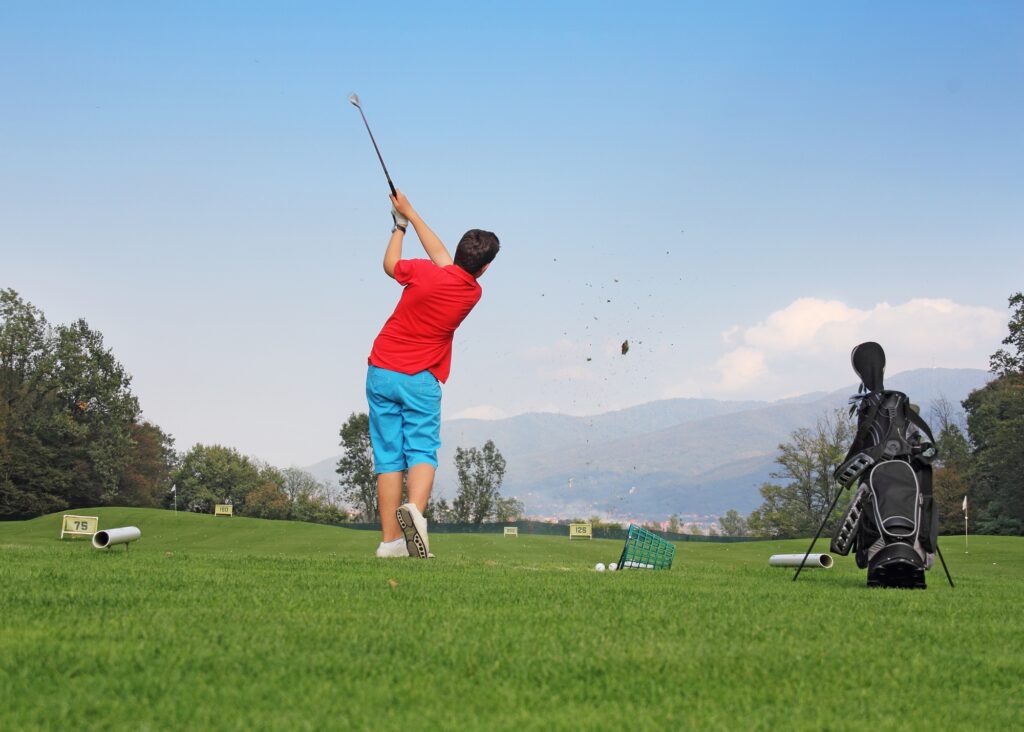 Golfer swinging at driving range with golf bag and practice balls on green field against mountain backdrop.